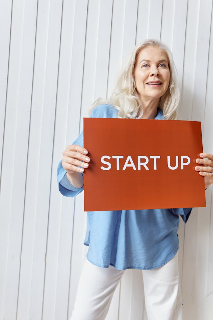 Elderly woman smiling while holding a 'Start Up' sign indoors, symbolizing entrepreneurship.